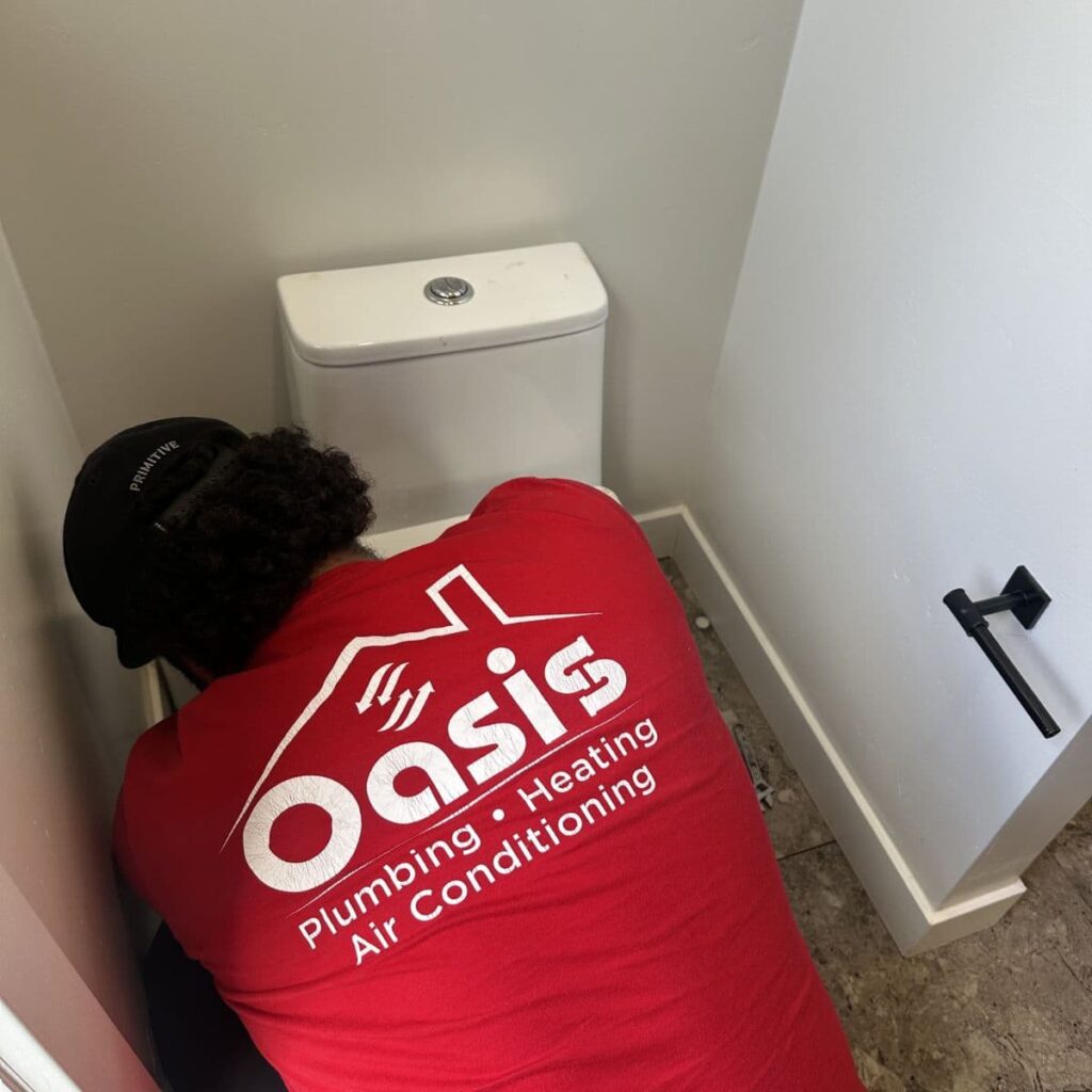 Plumber in an Oasis Plumbing & HVAC shirt servicing the back of a toilet in a compact bathroom