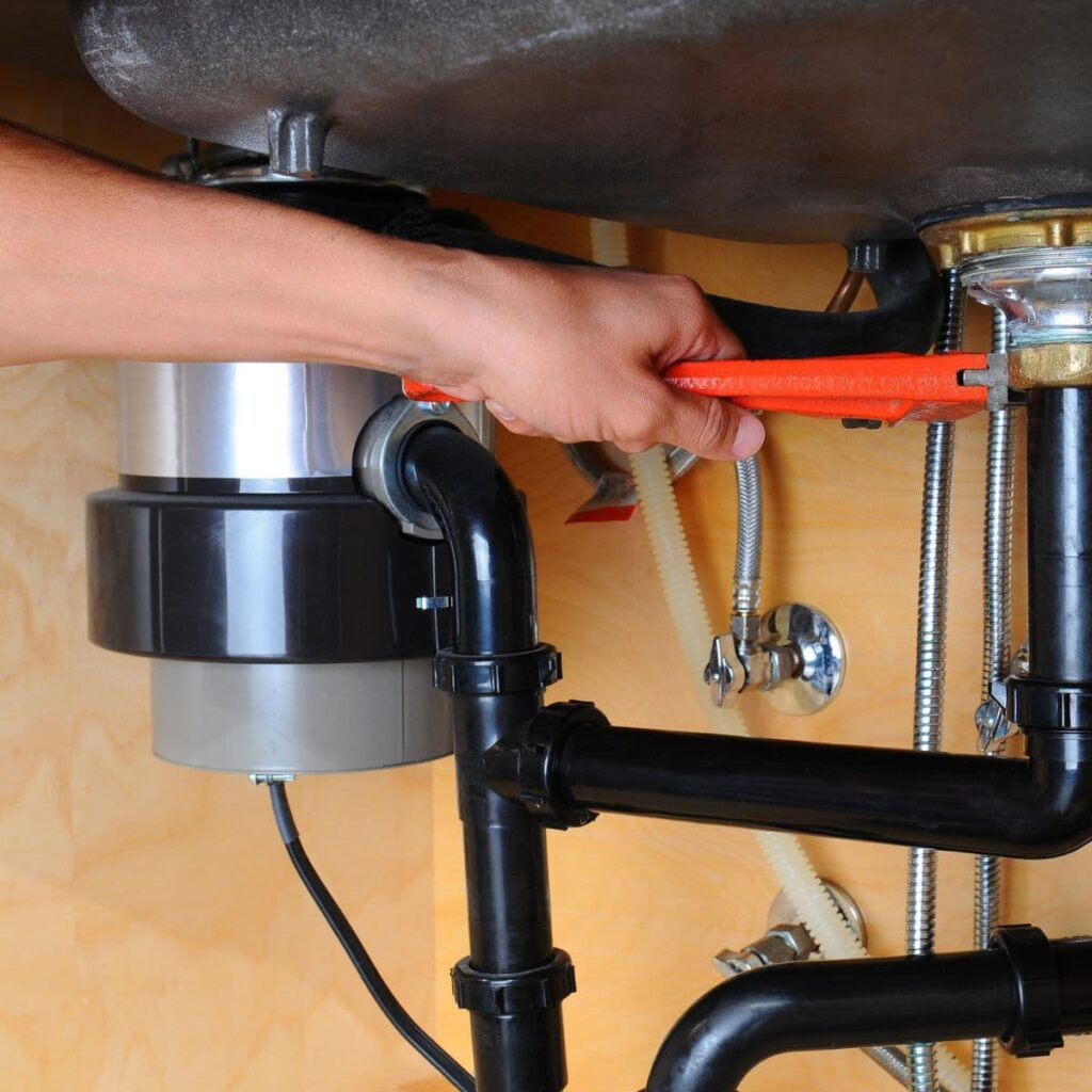 Plumber tightening black PVC pipes under a kitchen sink with a garbage disposal unit