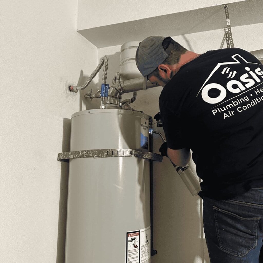Technician in Oasis Plumbing & HVAC shirt working on a tall, tank-style water heater with metal venting and supply lines