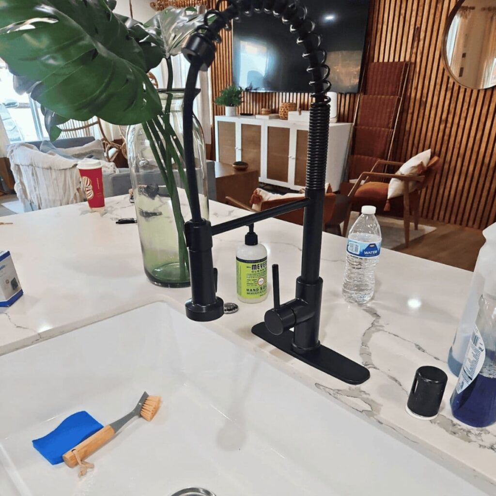 Matte black pull-down kitchen faucet with surrounding items like hand soap, scrub brush, and water bottle on a marble countertop