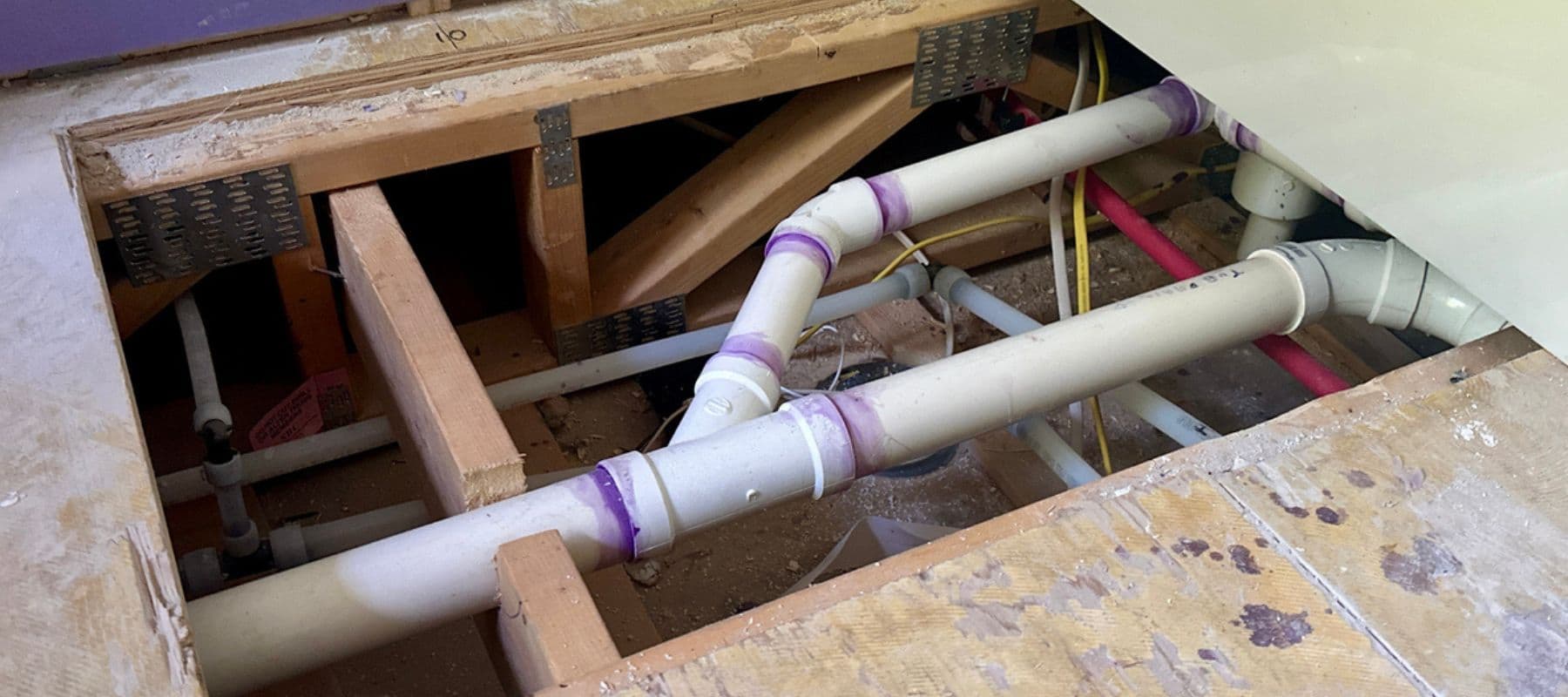 View of newly installed PVC plumbing lines under a wooden floor with purple primer marks