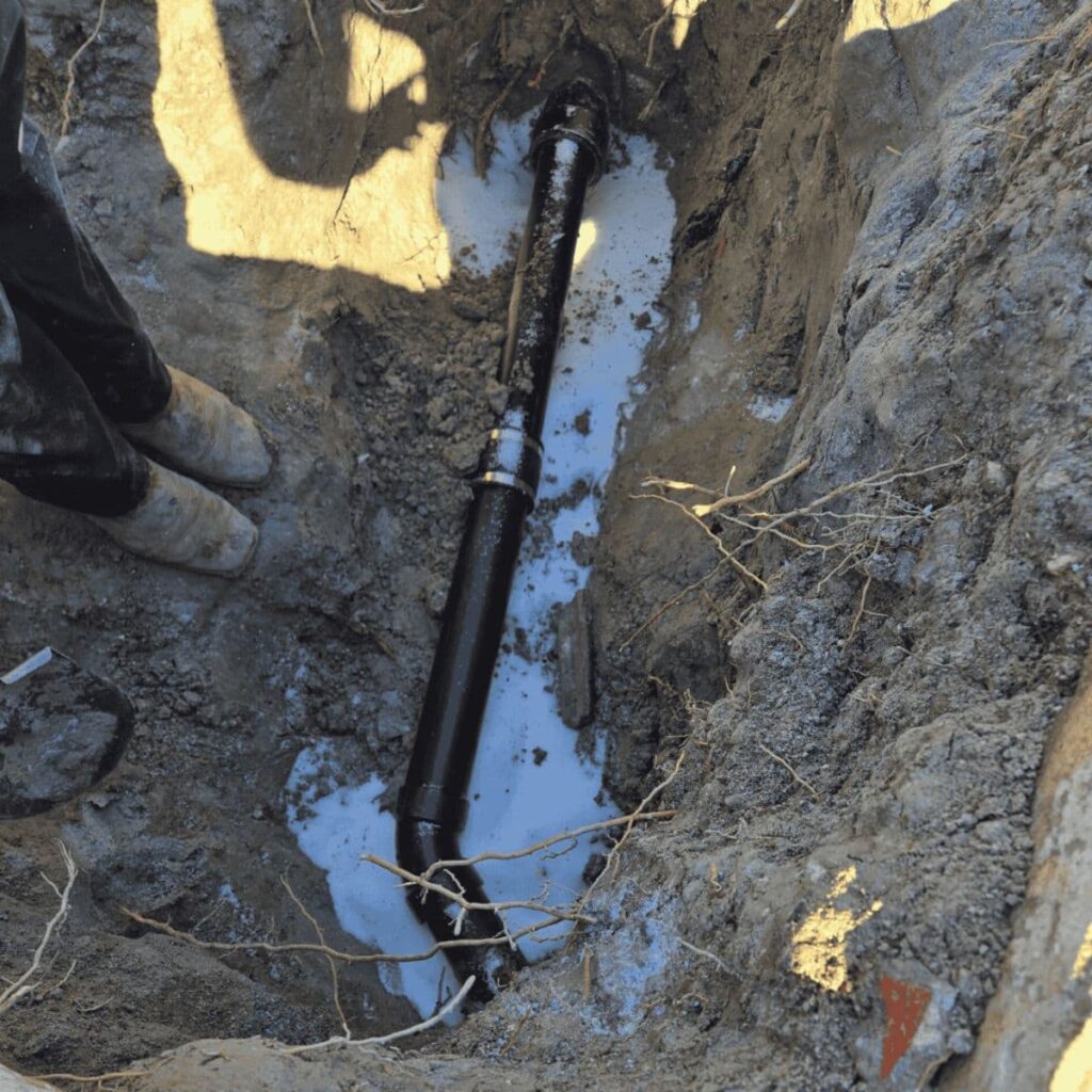 Black ABS sewer line partially buried in a dirt trench during underground plumbing work
